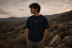 Man standing in a desert landscape wearing a black t-shirt with 'HERE' printed on it.