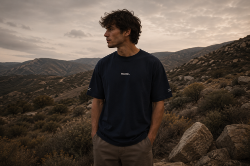 Man standing in a desert landscape wearing a black t-shirt with 'HERE' printed on it.