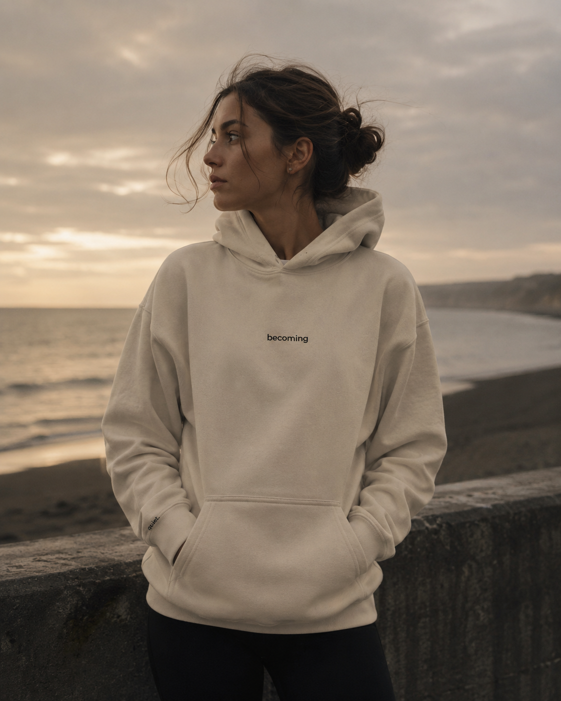 Person wearing a beige hoodie with 'becoming' text, standing on a beach at sunset.