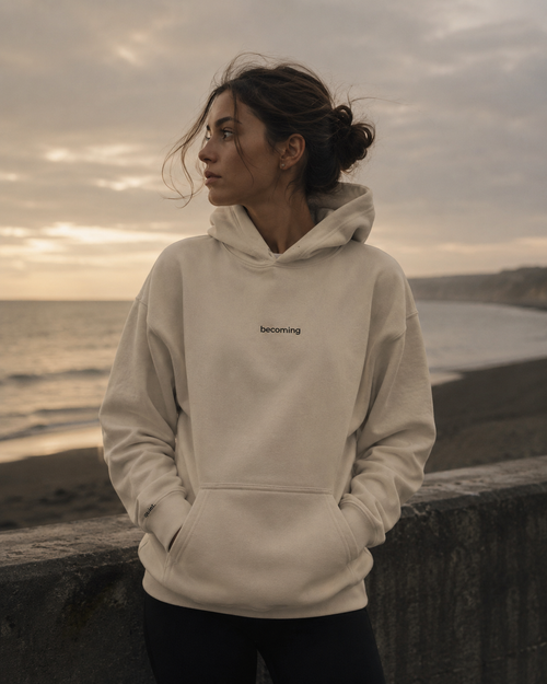 Person wearing a beige hoodie with 'becoming' text, standing on a beach at sunset.